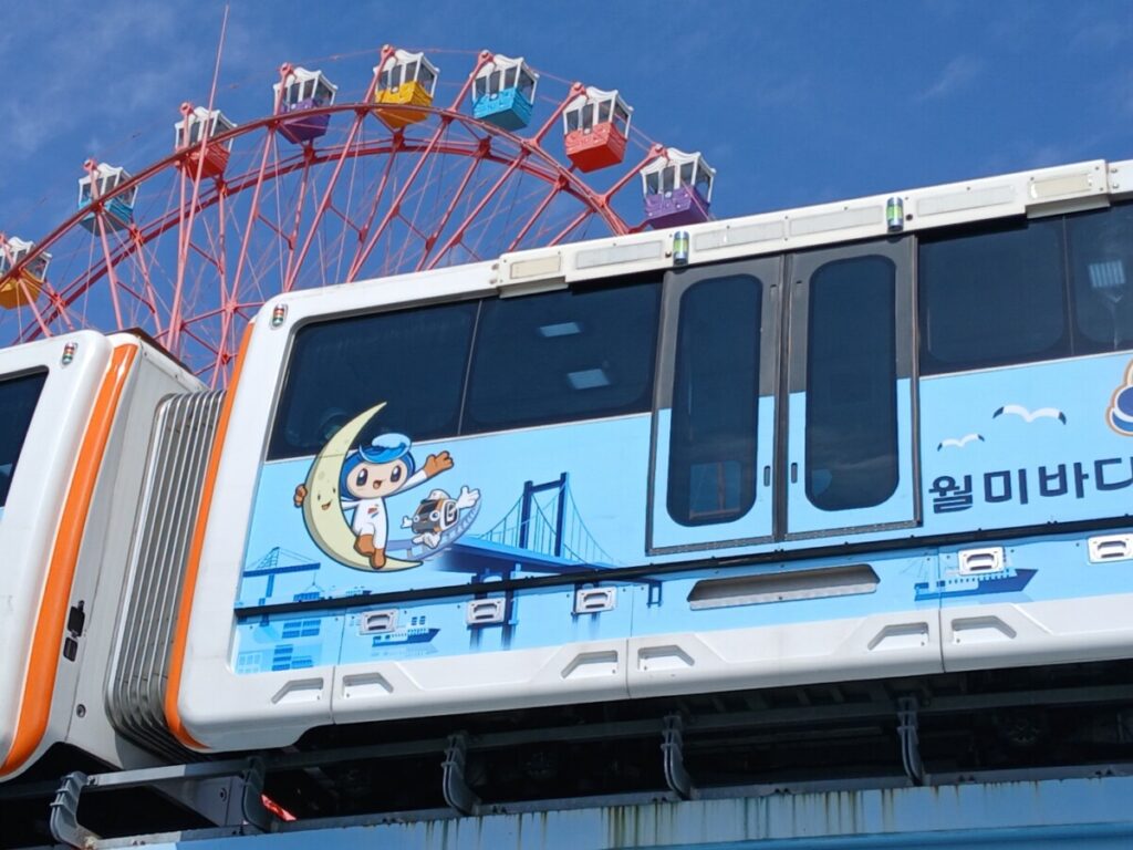 맑은 하늘 위 궤도를 따라 이동하는 인천 월미바다열차의 모습- 인천 월미바다열차를 타고 가며 창밖으로 흐드러진 벚꽃과 인천 내항의 풍경을 한눈에 담을 수 있는 특별한 경험을 선사합니다.